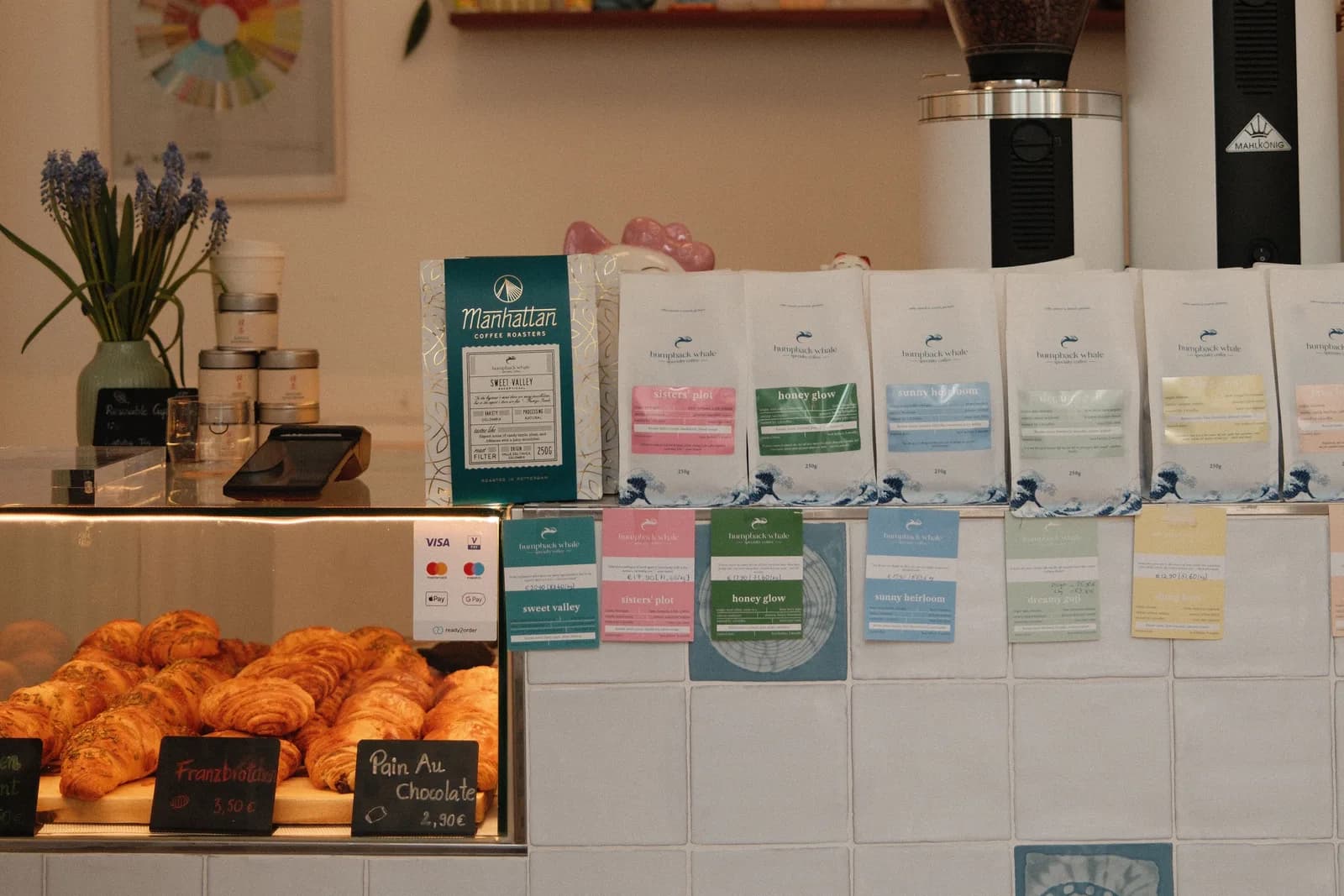 Die Theke des Humpback Whale Cafés in der Maxvorstadt — Croissants in der Vitrine, Kaffeemühlen und eine Reihe Single-Origin-Kaffeetüten, München