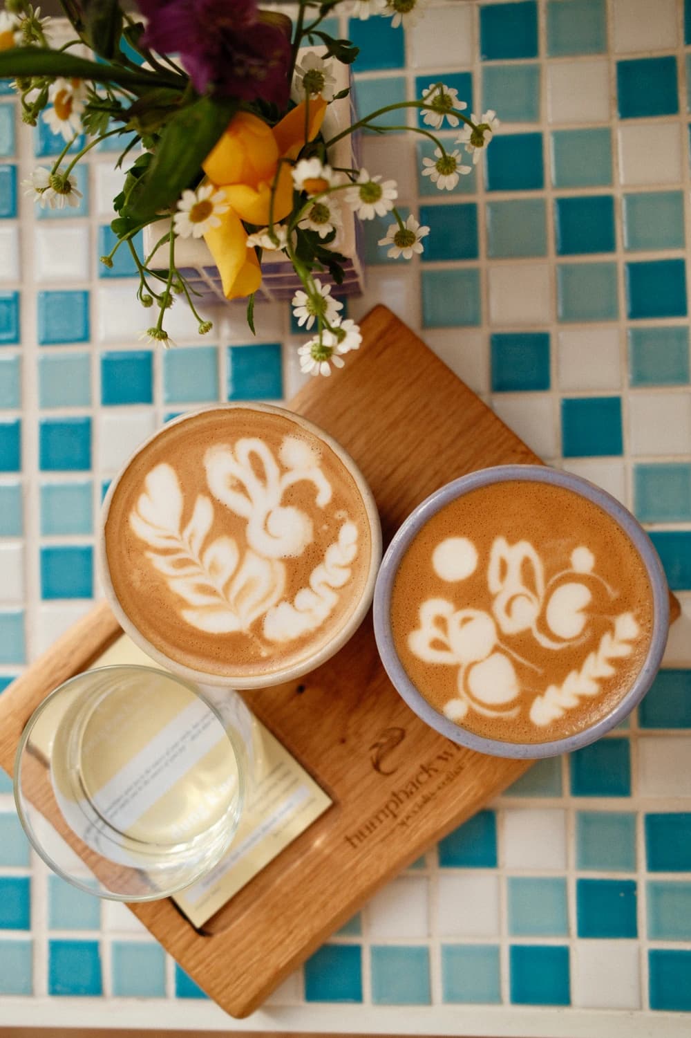 Zwei Flat Whites mit floraler Latte-Art in handgefertigten Keramiktassen auf einem Humpback-Whale-Holzbrett, München